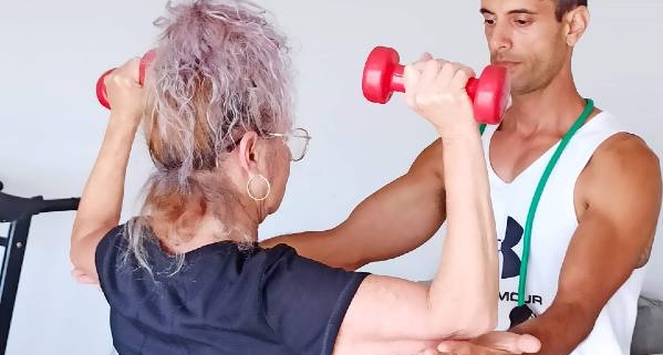 יוסי מאמן הכושר בגופיה לבנה מנחה מתאמנת האוחזת במשקולות אדומות