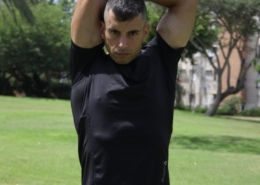 גבר בפארק בחולצת אימון שחורה אוחז יד מאחורי הראש במהלך שנראה כמו חימום לפני שמתאמנים