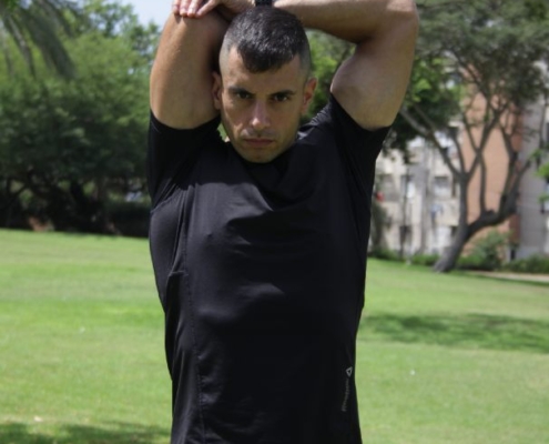 גבר בפארק בחולצת אימון שחורה אוחז יד מאחורי הראש במהלך שנראה כמו חימום לפני שמתאמנים
